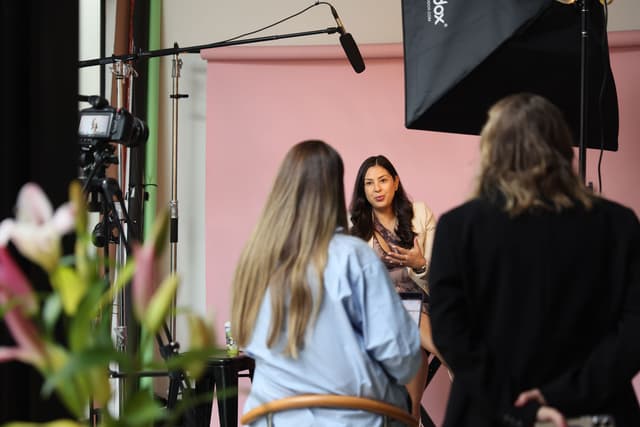 A woman speaks during an on-camera interview against a pink backdrop as crew members and a camera operator work around her.