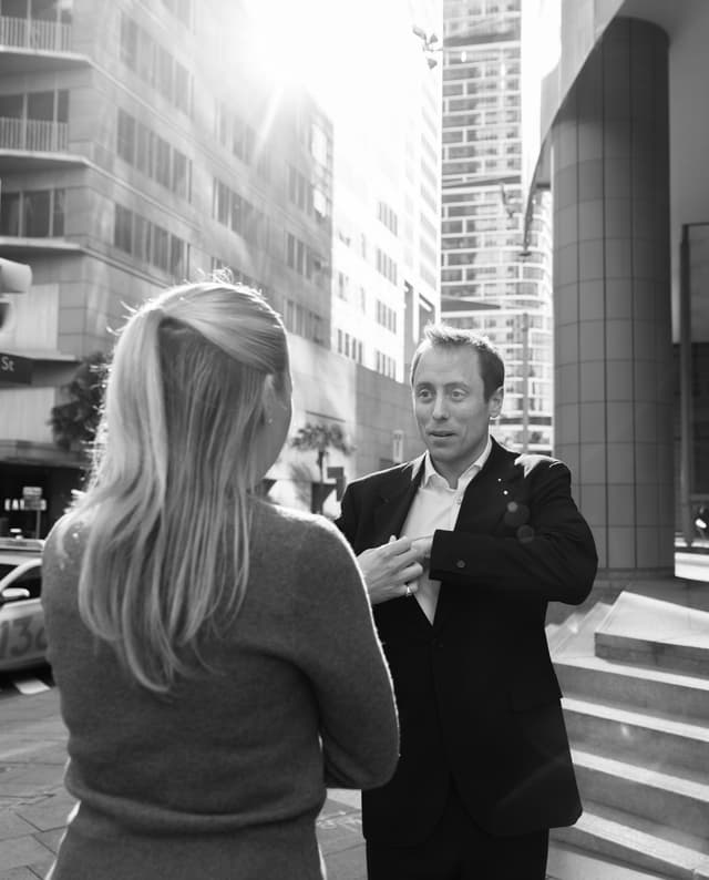 A man in a blazer speaks with a woman on a sunlit city street, captured in black and white.