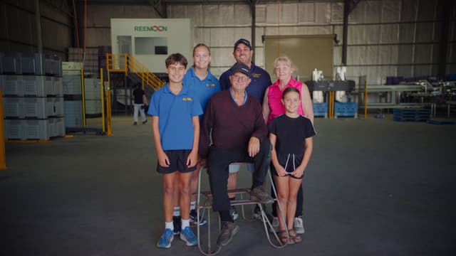 A family and staff pose together inside a Reemoon packaging warehouse with machinery and stacked crates.