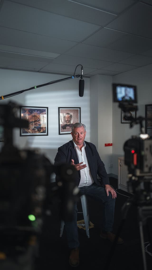 A man is interviewed on a stool in an office, framed between video camera equipment in the foreground.