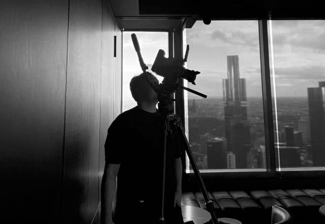 A silhouetted camera operator films a city skyline through a high-rise window, in black and white.