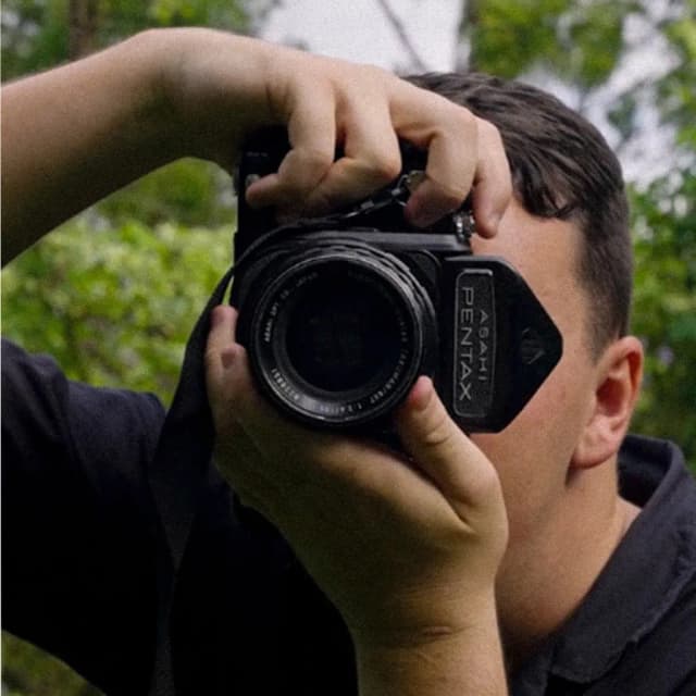 A man raises a vintage Pentax camera to his eye to take a photo outdoors.
