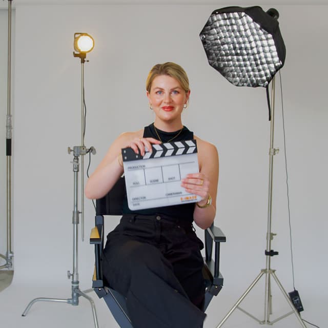 Karleigh Pearson sits in a director's chair holding a clapperboard between studio lights.
