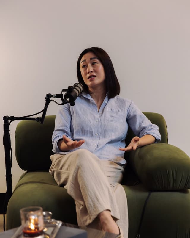 A woman speaks into a microphone while seated in a green armchair during a podcast recording for Is This Marketing.