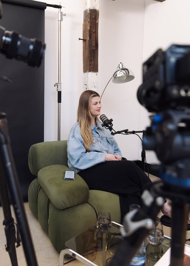 A woman is interviewed on a green couch surrounded by cameras and lighting equipment in a studio.