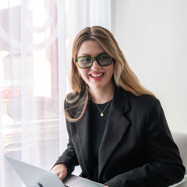 A woman wearing green glasses smiles while working on a laptop beside a sheer curtained window.
