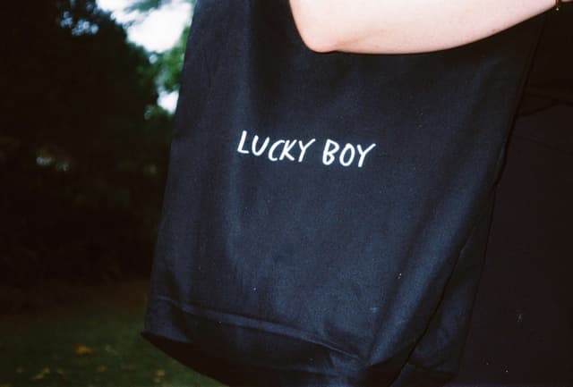 A close-up of a black tote bag printed with the words Lucky Boy, carried over someone's shoulder outdoors.