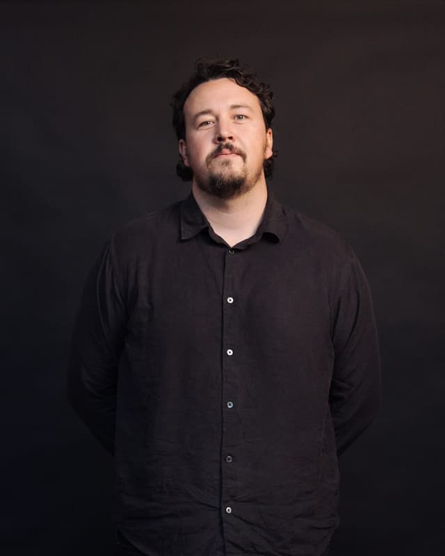 A bearded man in a black shirt poses for a studio portrait against a dark background.