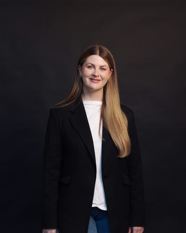 A studio portrait of a woman in a black blazer smiling against a dark background.