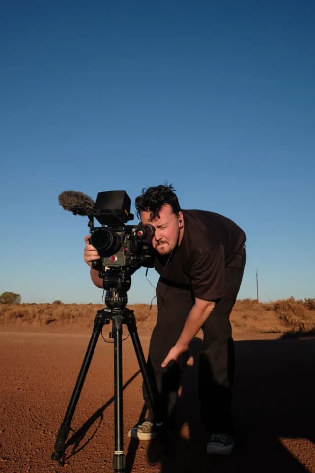 A cinematographer looks through a camera viewfinder on a tripod set up on a red dirt outback road.