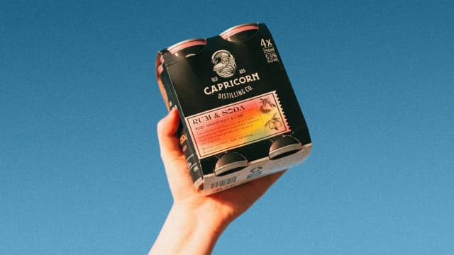 A hand holds up a four pack of Capricorn rum and soda cans against a clear blue sky.