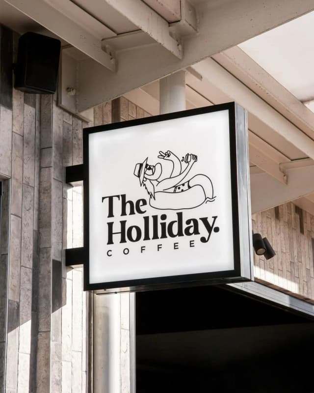 An illuminated shopfront sign for The Holliday Coffee featuring an illustrated character holding a drink.