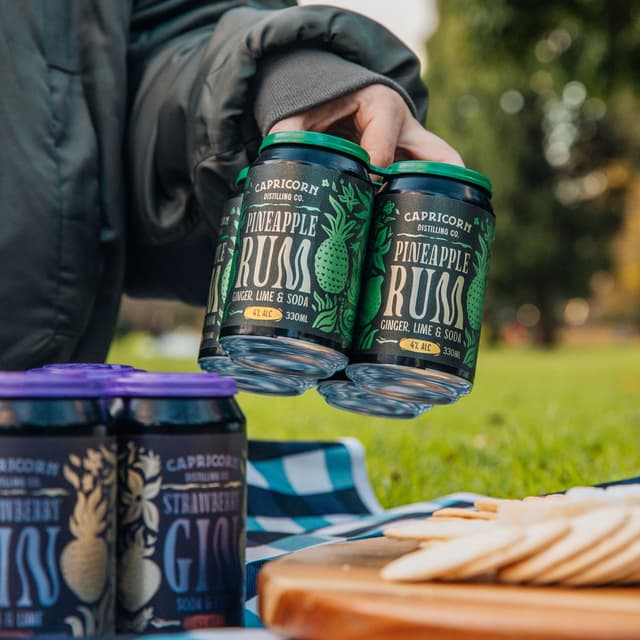 A hand holds a four pack of Capricorn Distilling Co pineapple rum cans over a picnic spread.
