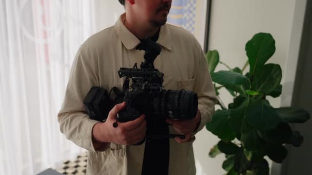 A man holds a cinema camera fitted with a zoom lens in a bright studio.