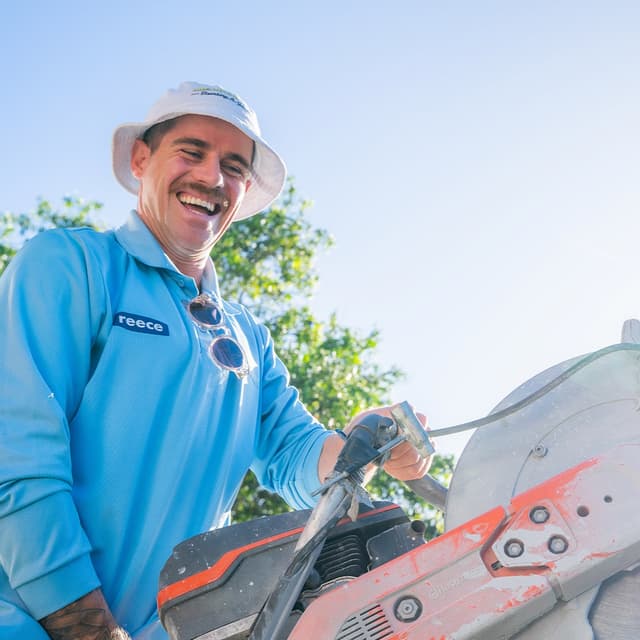 A smiling tradesman in a reece branded shirt holds a petrol cut off saw outdoors under a blue sky.