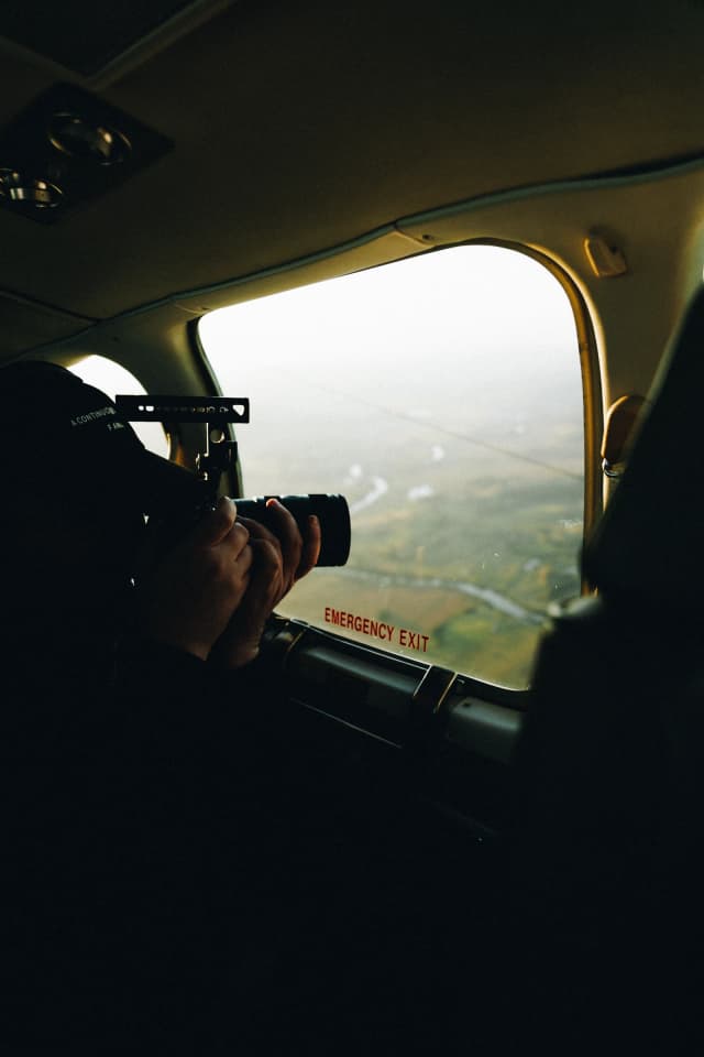 A photographer aims a camera out a small plane's window marked emergency exit to capture the landscape below.