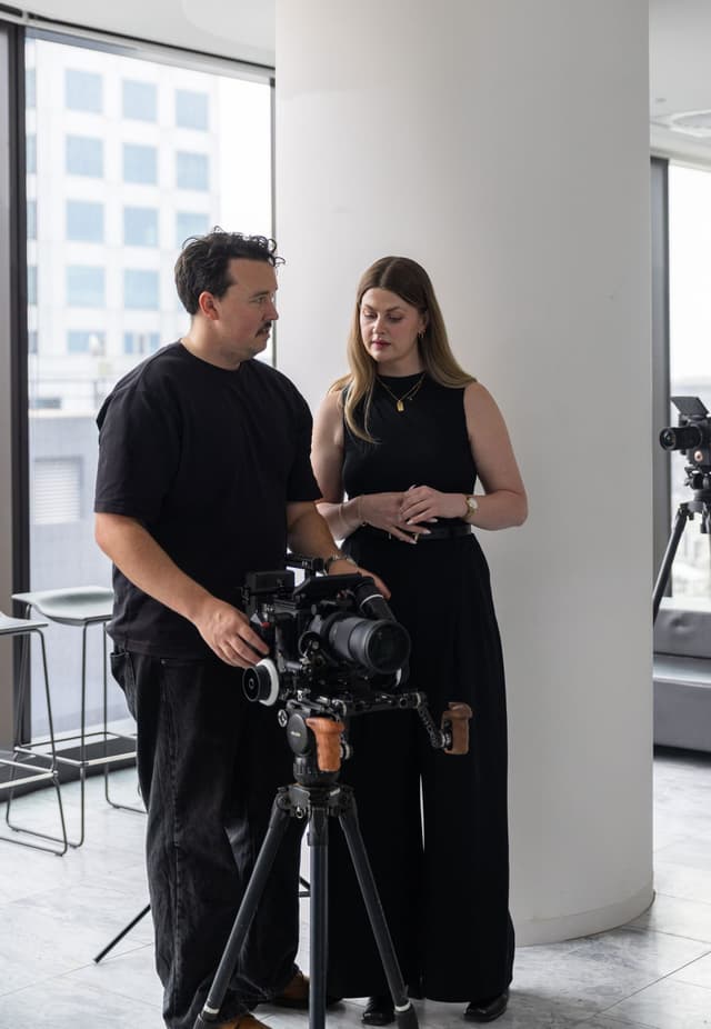 A camera operator and a colleague review footage on a cinema camera rig in a city office.