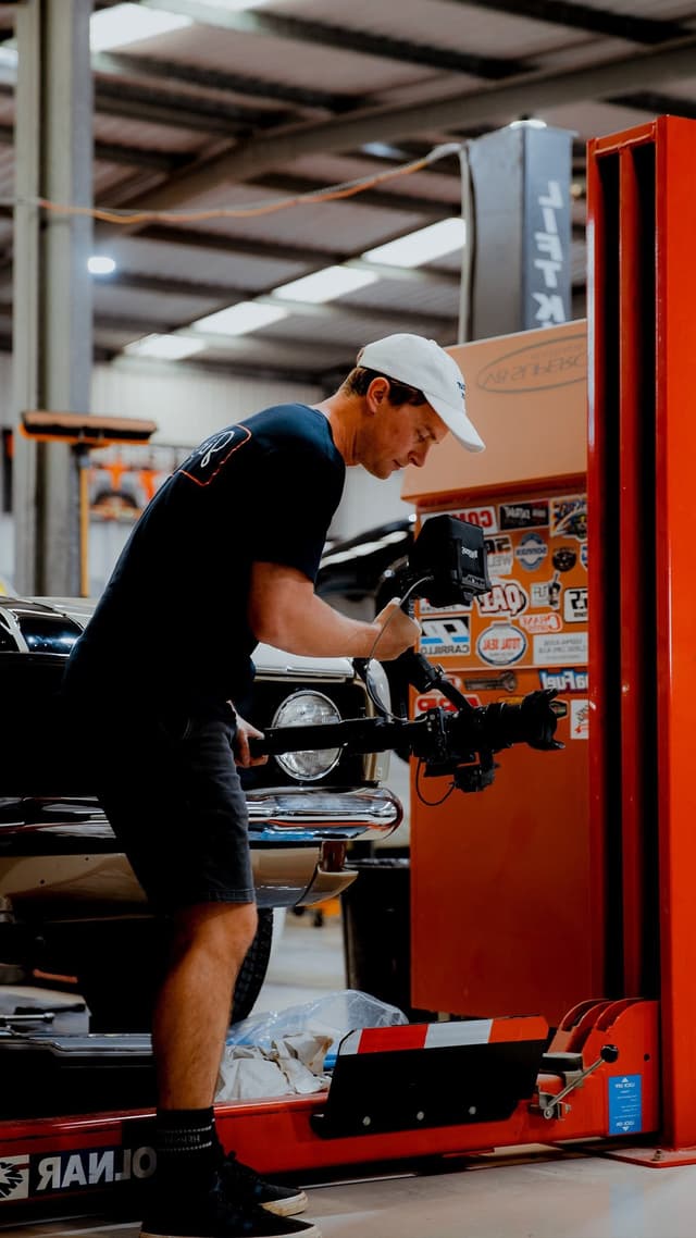 A man wearing a white cap films a classic car's engine bay with a camera rig in a sticker-covered workshop.