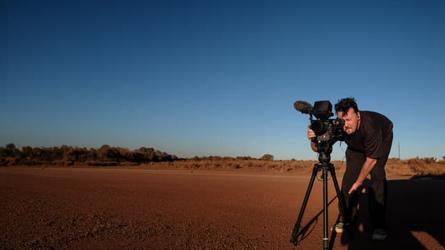 A camera operator leans into the viewfinder of a cinema camera on a tripod, on red outback dirt under a clear blue sky.