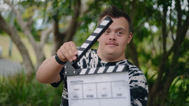 A young man holds a film clapperboard and smiles at the camera amid trees on an outdoor video shoot.