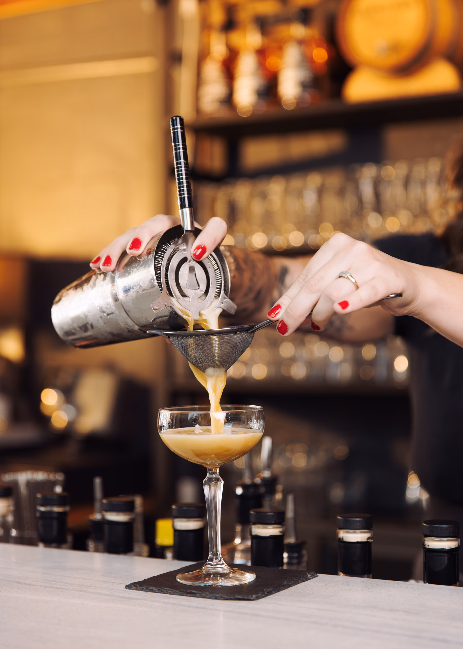A bartender strains a frothy cocktail from a shaker into a coupe glass on a Capricorn Distilling Co bar.