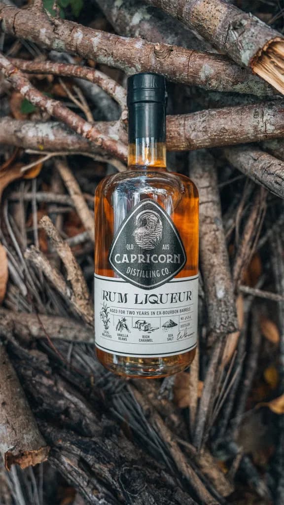A bottle of Capricorn Distilling Co rum liqueur rests among bare branches and dry leaves.