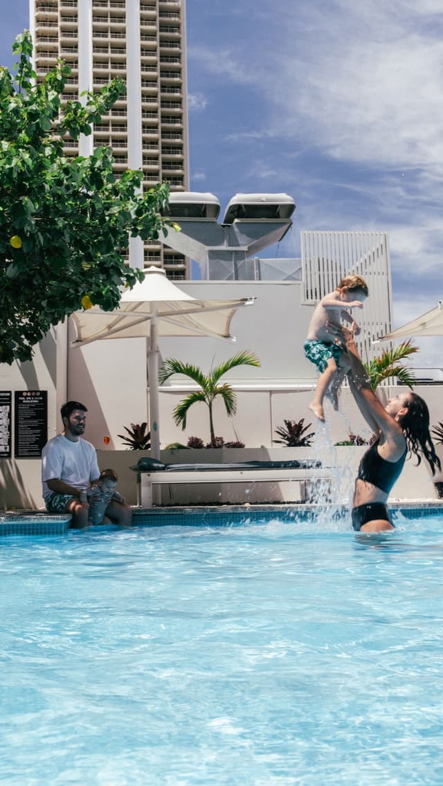 A mother tosses a laughing child in the air in the Hilton Surfers Paradise rooftop pool as family watches on.